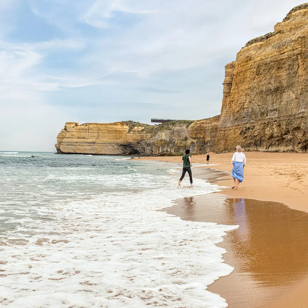 Two people walking along the water's edge on a sandy beach beneath tall golden limestone cliffs