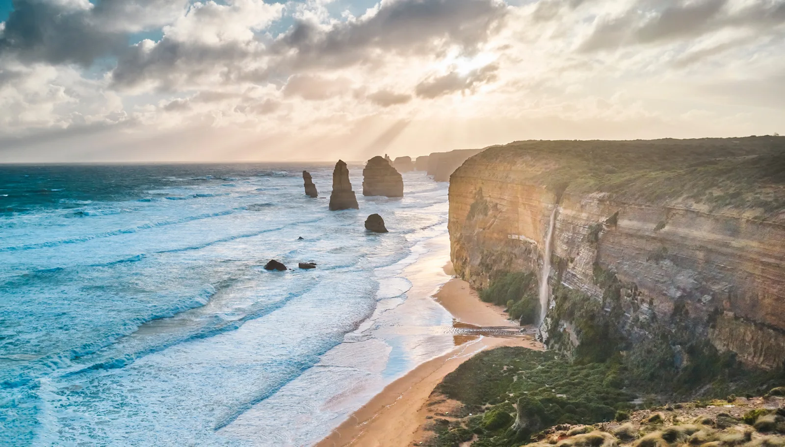 Sunlight breaking through clouds over the Twelve Apostles, with turquoise waves rolling onto the beach beneath tall limestone cliffs