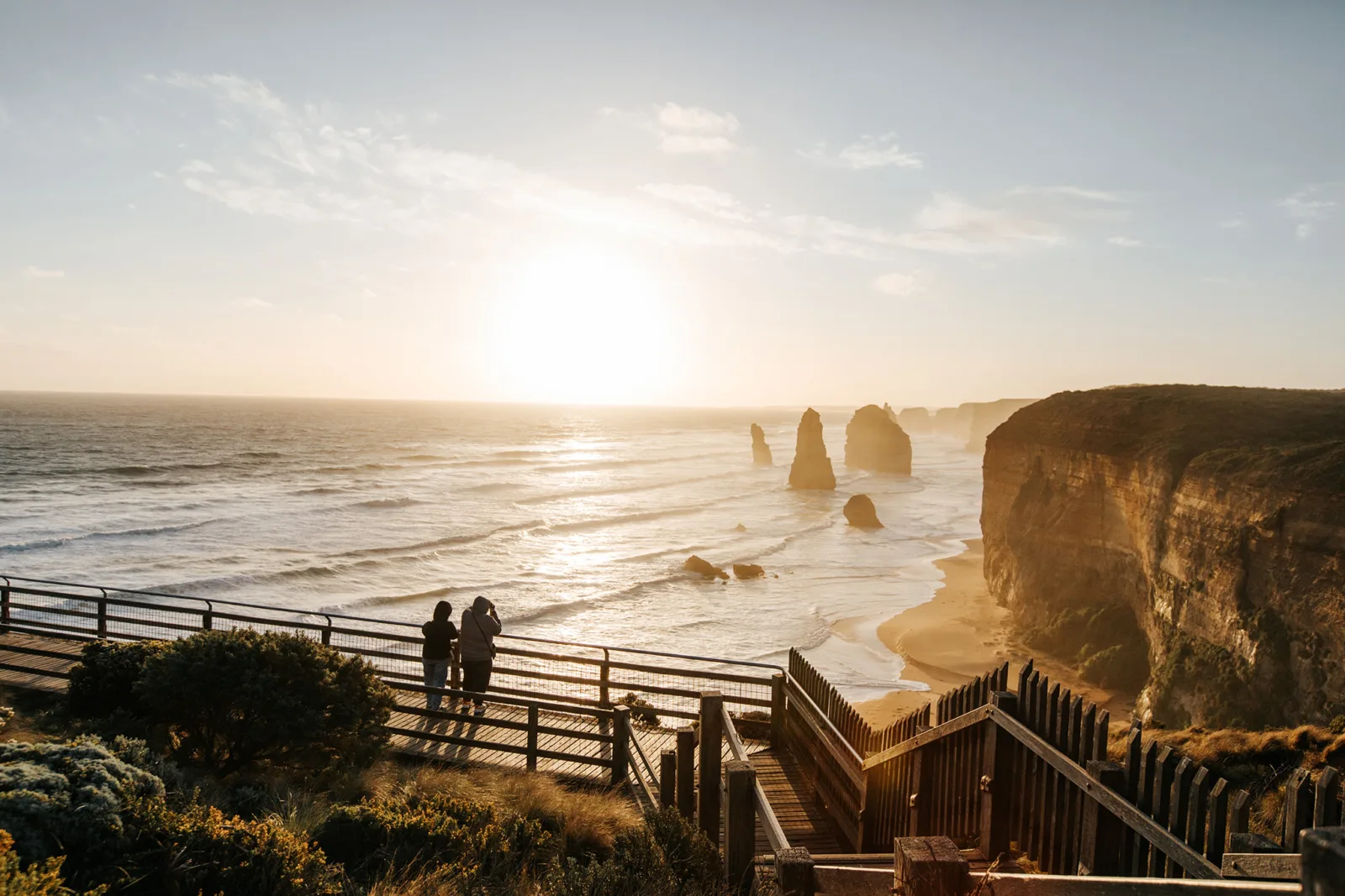 Two visitors on a timber viewing platform watching the sun set over the Twelve Apostles and the Southern Ocean