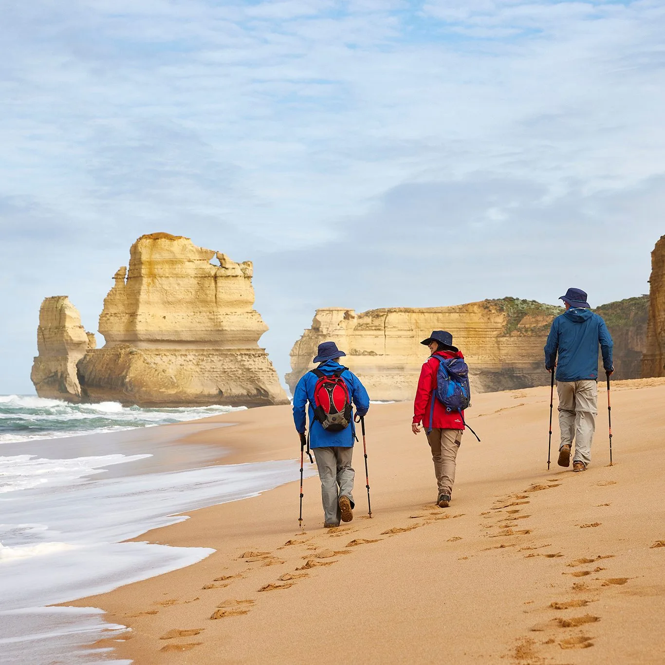 Three hikers with backpacks and trekking poles walking along a sandy beach toward the Twelve Apostles limestone stacks