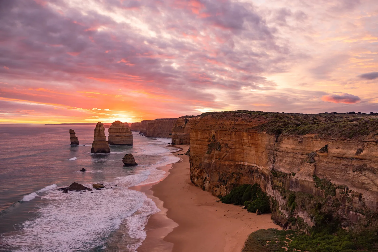 Pink and orange sunset clouds over the Twelve Apostles, with the sun setting on the horizon beside towering golden cliffs