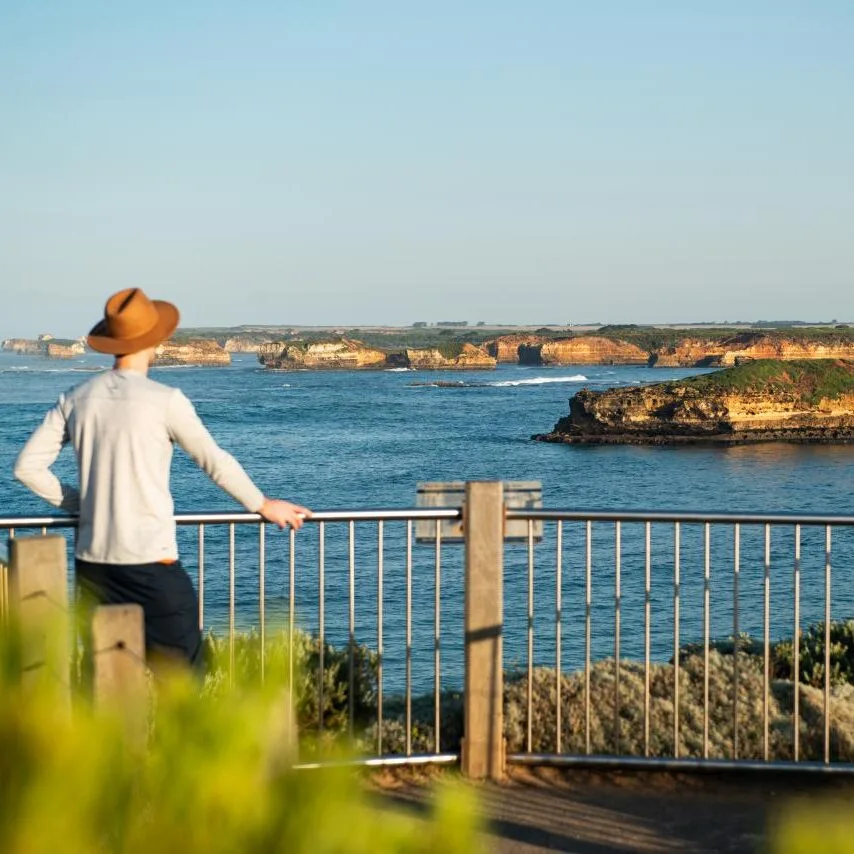 A visitor in a hat leaning on a lookout railing, gazing out over a calm bay dotted with rock formations