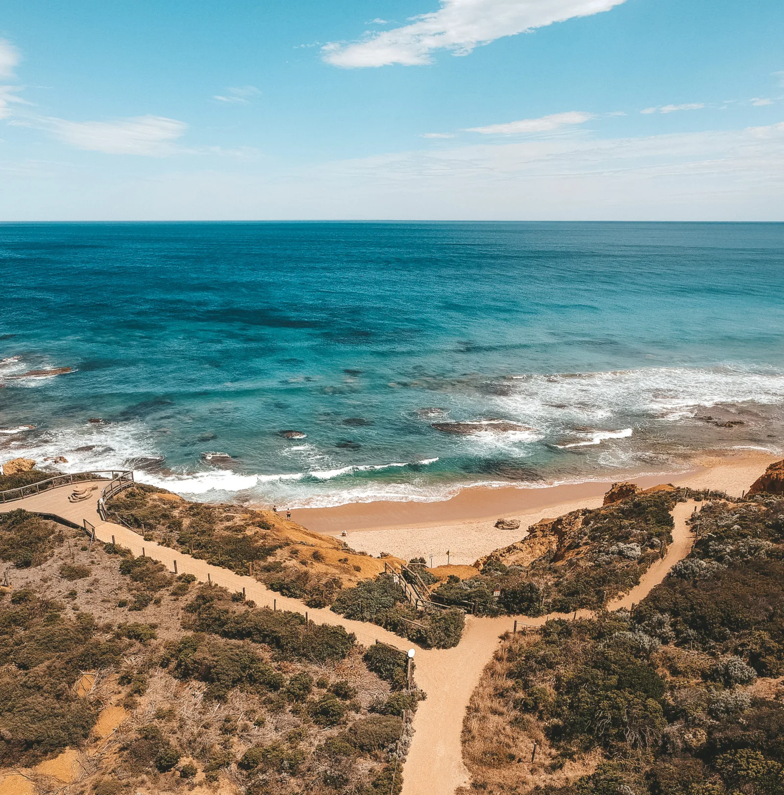 Sandy walking paths winding through coastal scrub down to a beach, with bright blue ocean and rolling surf beyond