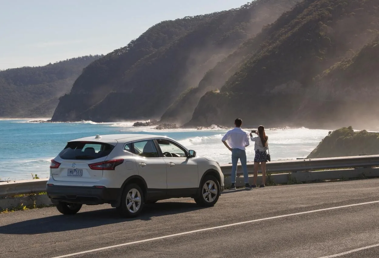 A couple standing beside their parked car at a roadside pull-off, taking in views of the ocean and steep coastal hills along the Great Ocean Road