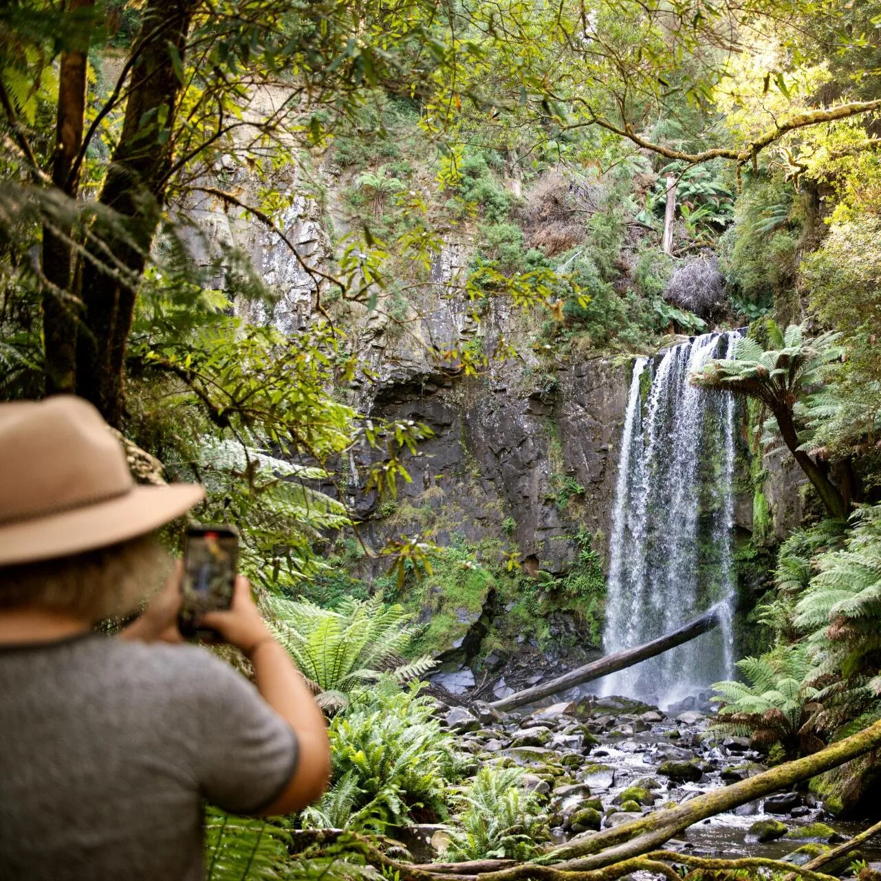 A visitor in a wide-brimmed hat photographing a waterfall cascading through lush green rainforest