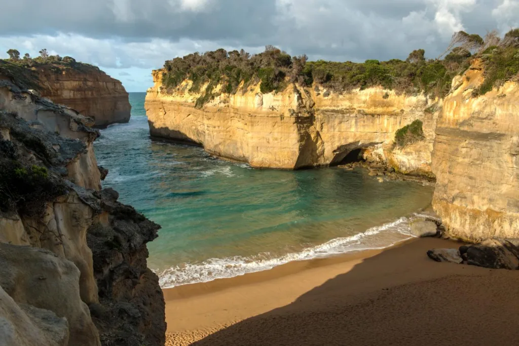 Loch Ard Gorge from above, with turquoise water lapping a sheltered sandy beach enclosed by sunlit limestone cliffs topped with coastal scrub