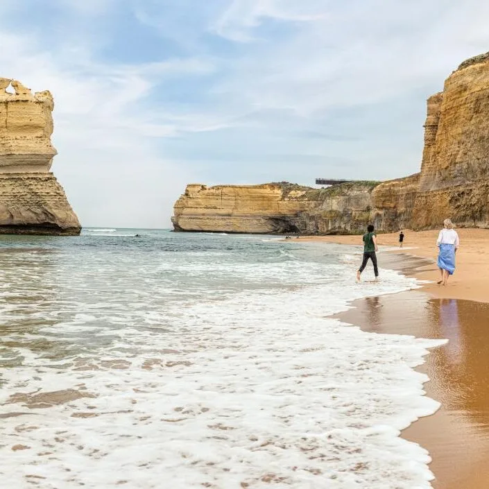 Two people strolling through the shallows beside limestone stacks and cliffs on a sunny day