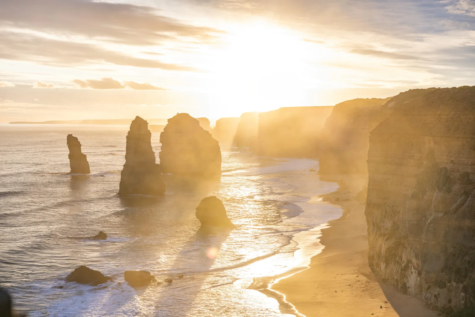 Bright golden sunlight streaming over the Twelve Apostles and coastal cliffs, casting the limestone stacks in silhouette