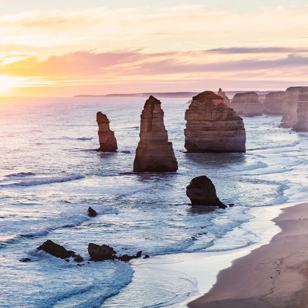 Twelve Apostles limestone stacks at sunset, with soft golden light over the ocean and waves washing onto the beach