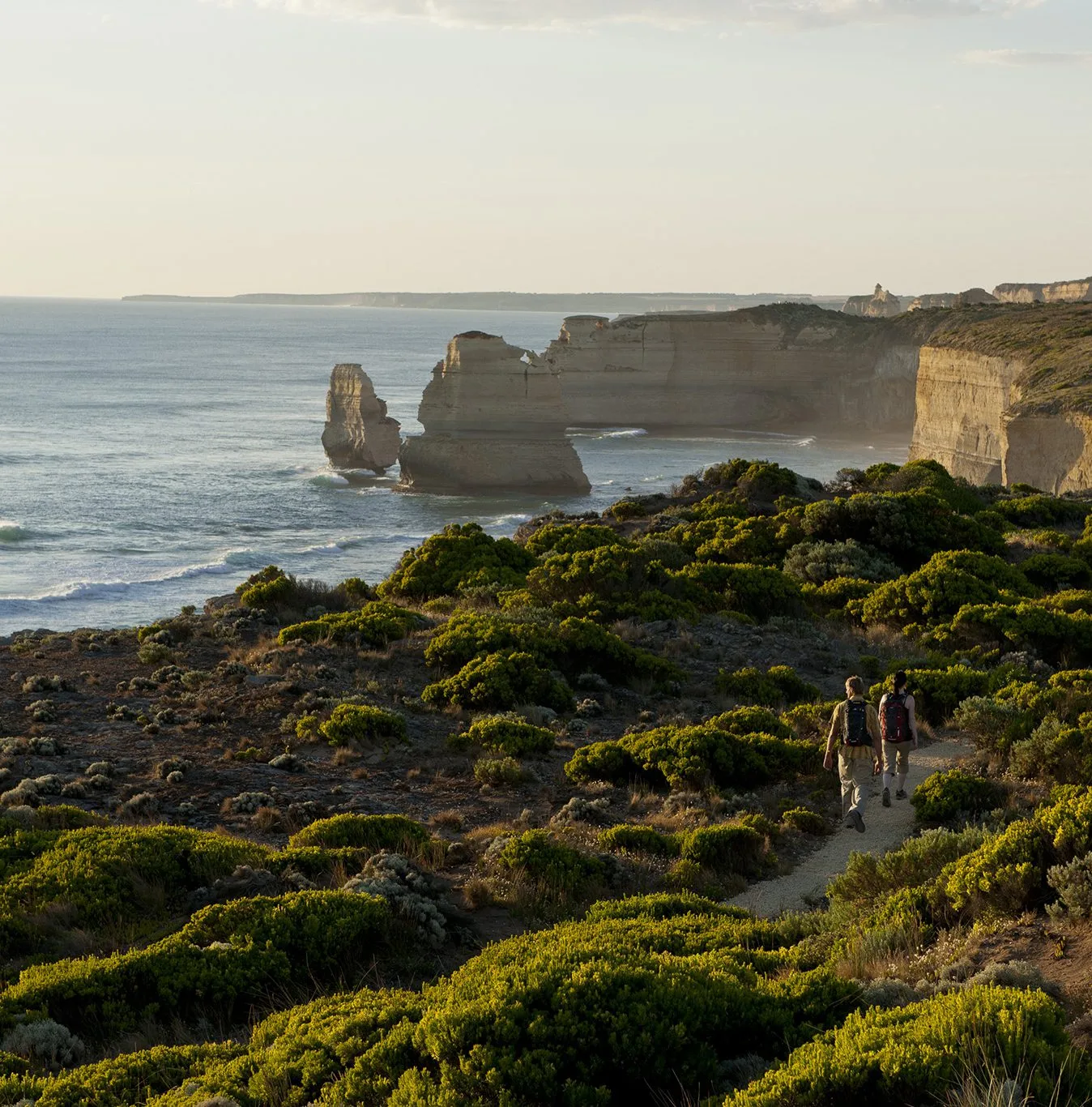 Two walkers on a trail through green coastal heath, heading toward limestone stacks and cliffs in late afternoon light