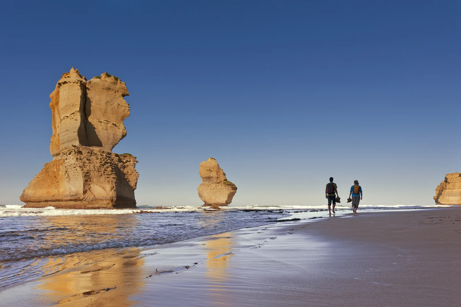 Two people walking along the wet sand toward a limestone stack, lit by warm golden light under a clear blue sky