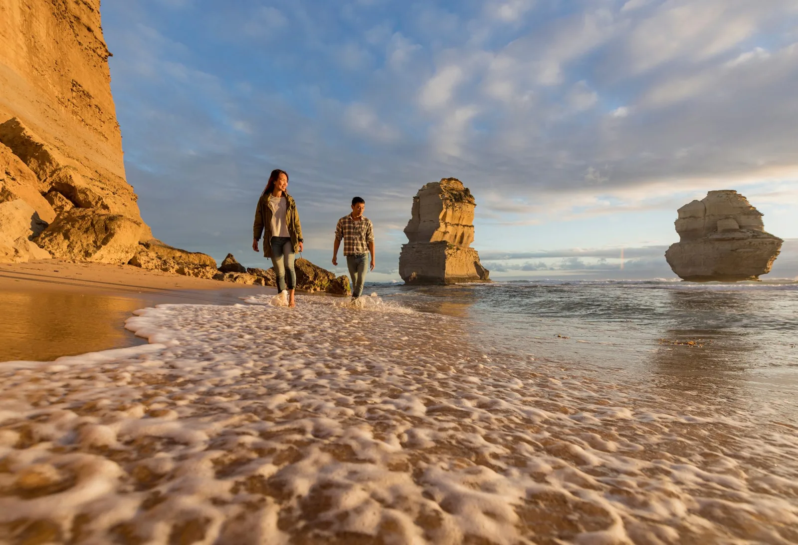 Two people walking barefoot along a sandy beach toward a limestone stack, with gentle waves and a blue sky overhead