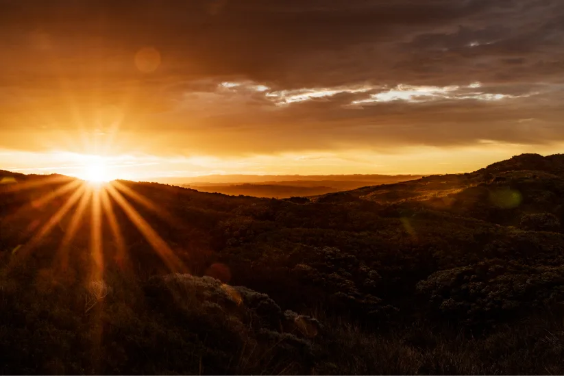 The sun setting over rolling coastal hills covered in dense bushland, with golden rays streaming across a moody, cloud-filled sky