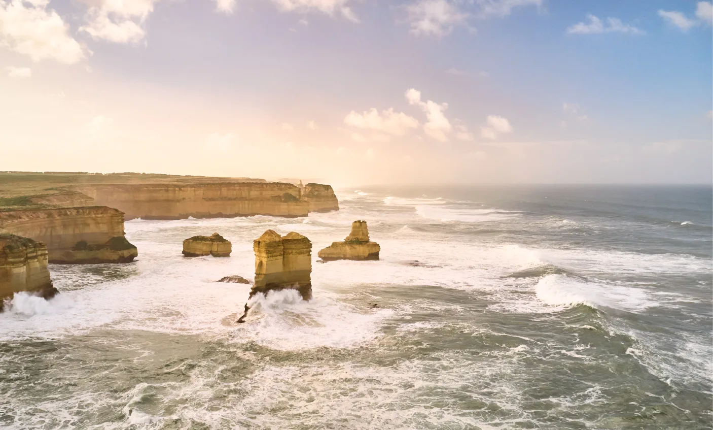 Aerial view of limestone stacks standing in rough white surf off the Twelve Apostles coastline, with golden cliffs glowing in hazy afternoon light
