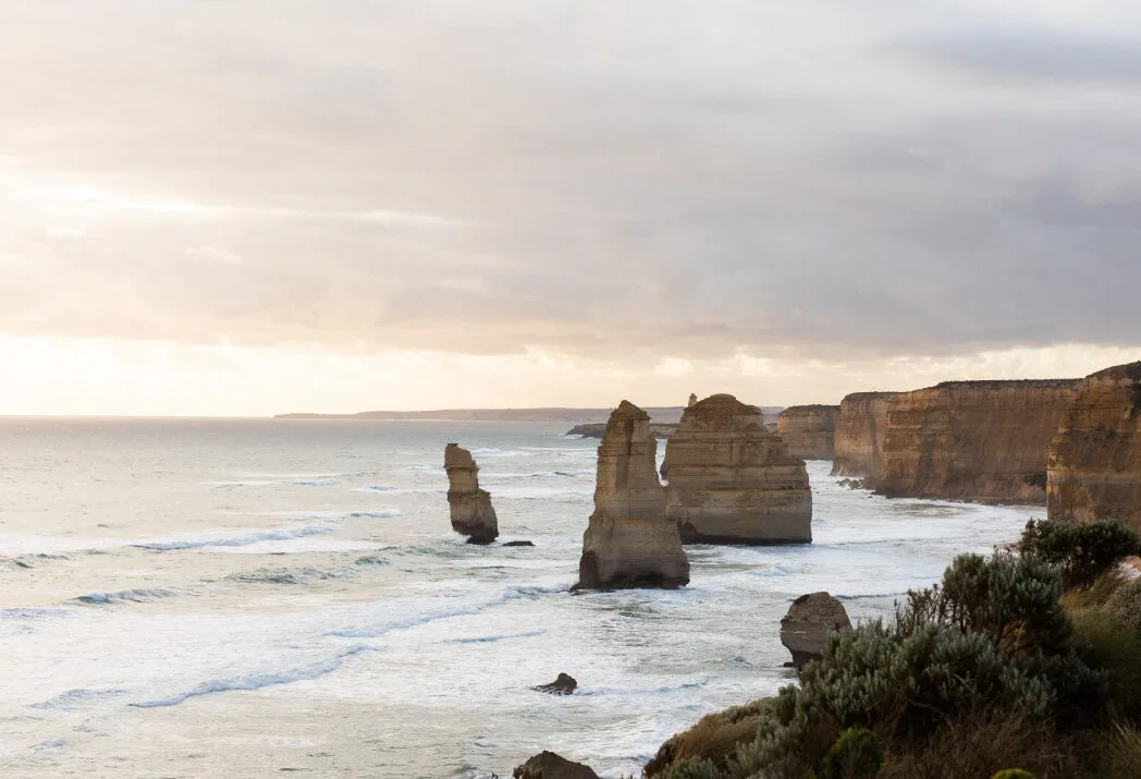 The Twelve Apostles under a soft, overcast sky, with gentle waves washing around the limestone stacks below the cliffs