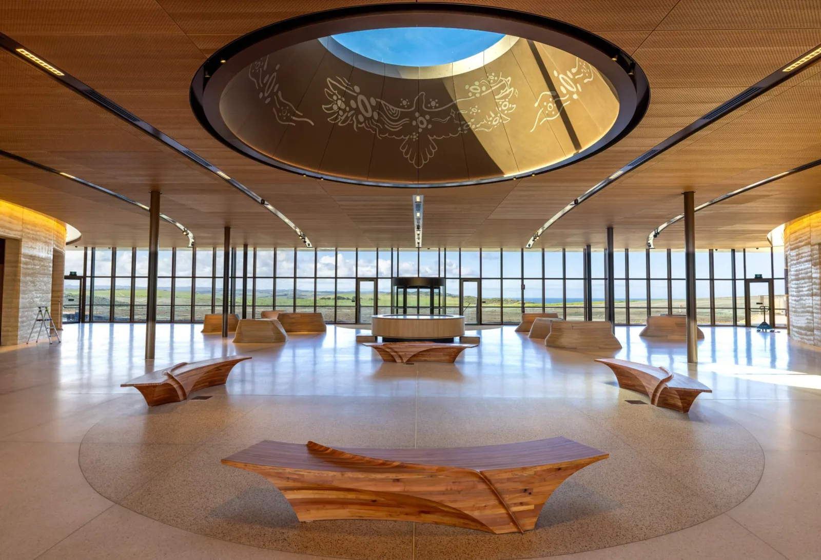 Sculpted timber benches arranged in a circle beneath the foyer's illuminated skylight, with floor-to-ceiling windows looking out over the landscape