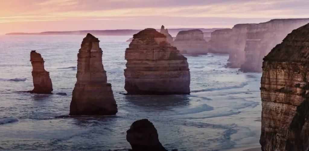 Close view of the Twelve Apostles limestone stacks rising from the sea, with misty cliffs fading into a soft pink and lilac sky