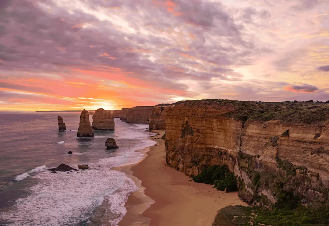 Twelve Apostles limestone stacks rising from the Southern Ocean at sunset, with golden cliffs and a sandy beach below a pink and purple sky